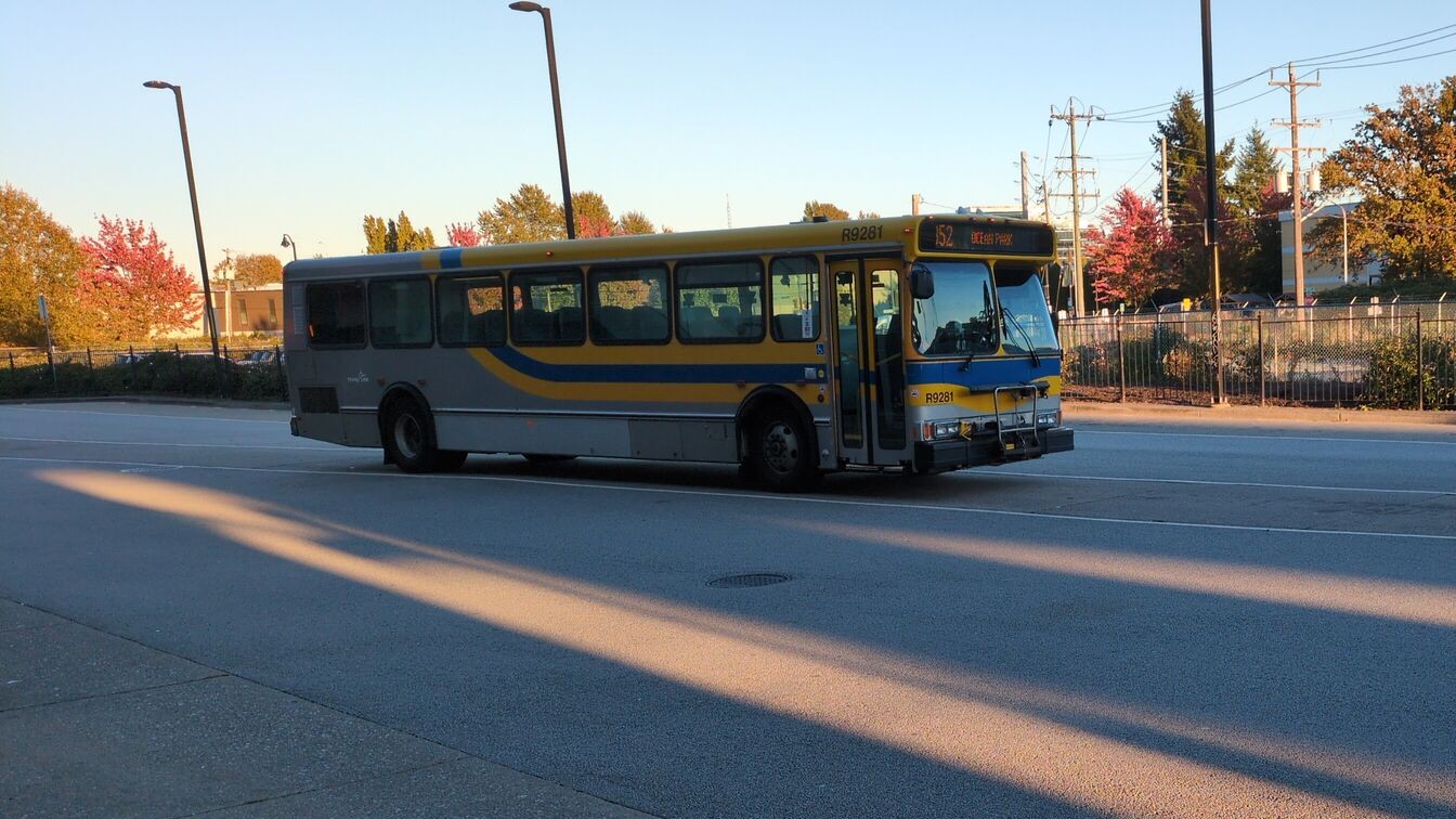 A bus with the CMBC suburban grey color scheme (Grey body, yellow stripe, blue stripe and another yellow strip going to the roof of the bus. the stripes curves upward towards the end of the bus.) is parked at a bus loop. The destination sign says '152 ocean park'